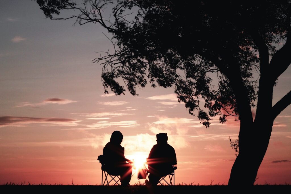Couple sat in camping chairs by a tree looking over a beautiful sunset.