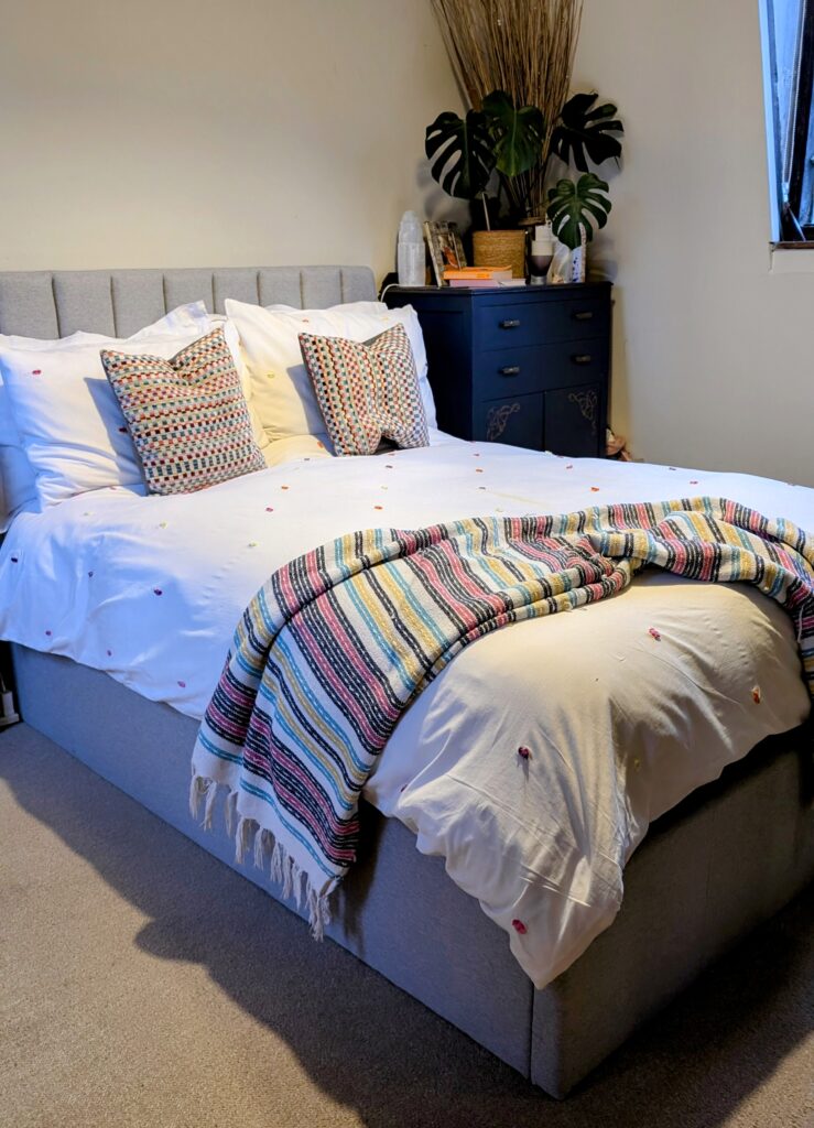 A grey bed frame with a white bed set it has coloured dogs and a rainbow coloured throw and cushions.